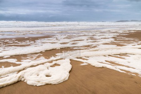 Photo for Stormy sea and skies and a beach covered in sea foam - Royalty Free Image