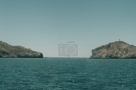 Calm seascape, sailboats near rocky coastline under blue sky