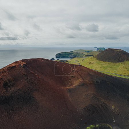 Aerial view of the volcanic landscape in Vestmannaeyjar (Westman Islands), Iceland, showcasing the rugged terrain, red volcanic hills, and distant islands set against the expansive ocean. 
