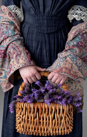 Eine junge viktorianische Frau trägt ein blaues Baumwollkleid mit Spitzenbesatz und hält einen Korb Lavendel vor einer Studiokulisse