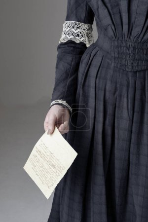Eine junge viktorianische Frau trägt ein blaues Baumwollkleid mit Spitzenbesatz und hält einen Brief vor einer Studiokulisse