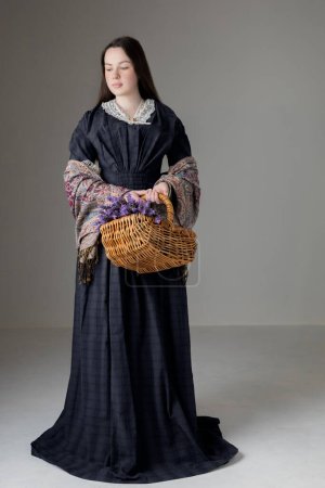 Une jeune femme victorienne portant une robe en coton bleu avec des garnitures en dentelle vintage et tenant un panier de lavande sur fond de studio