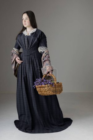 A young Victorian woman wearing a blue cotton dress with vintage lace trim and holding a basket of lavender against a studio backdrop