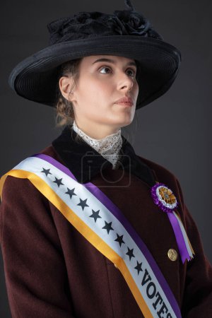 American Victorian or Edwardian Suffragette wearing historically accurate purple and gold colored sash and rosette against a studio backdrop 