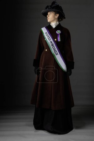 Reino Unido Victoriana o Edwardian Suffragette vistiendo históricamente precisa banda púrpura y verde y roseta y protestando por los derechos de voto de las mujeres en un fondo de estudio