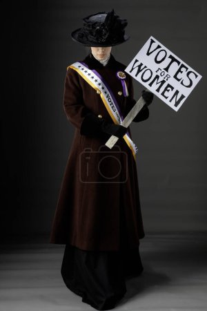 Sufragista estadounidense victoriana o eduardiana que viste el marco y la roseta púrpura y dorada históricamente precisos y protesta por los derechos de voto de las mujeres contra un telón de fondo de estudio llano