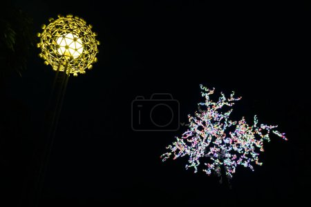 Una escena nocturna con una lámpara de calle brillante y decorativa a la izquierda y un árbol adornado con luces de colores a la derecha.