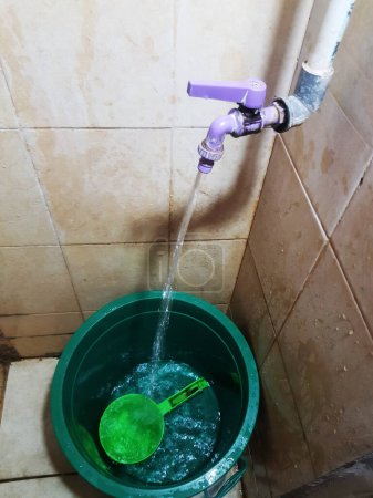 Water flowing from a faucet into a green bucket with a green ladle, in a tiled bathroom.