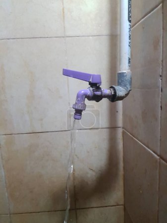 Water flowing from a purple tap against a tiled wall, showing signs of wear and mineral deposits