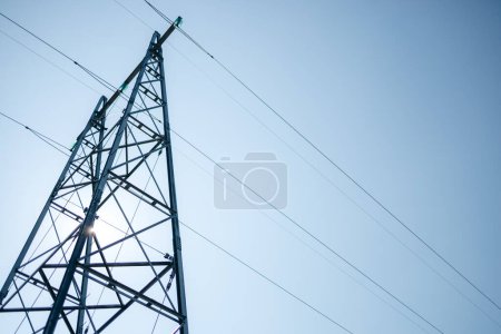 Frogs eye view of an electrical mast and cables, with the sun hidden behind it on blue, clear sky. Field in Norway.