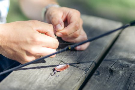 Closeup of hands tying a fishing knot on a fishing lure on a wooden table with fishing rod in sunshine. Summer in Norway.