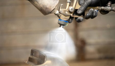 Imagen de cerca de las manos con guantes de un trabajador calificado usando una pistola de pulverización profesional para pintar una pieza de automóvil, destacando el pulverizador de precisión y la niebla fina.