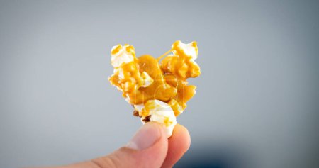Eine Person hält eine Traube karamellbeschichteten Popcorn in der Hand, das vor einem gedämpften blauen Hintergrund präsentiert wird und die Textur und goldene Farbe des Snacks betont..