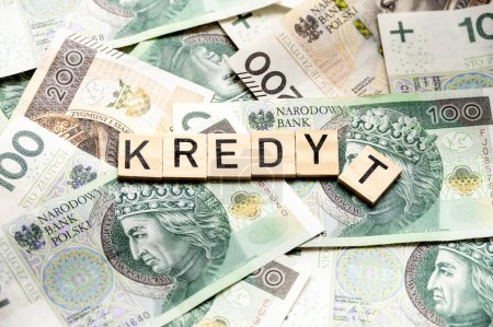 Une image visuellement engageante capturant des blocs de bois orthographiant 'Kredyt', entourée de diverses dénominations de billets de banque polonais en zloty. Il met l'accent sur les thèmes de la finance, de la dette et de la banque en Pologne.