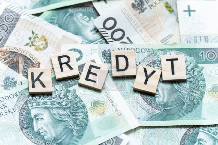 L'image montre des carreaux de Scrabble en bois orthographiant le mot polonais 'KREDYT', qui signifie crédit, disposés sur une pile de monnaie zloty polonaise, symbolisant des thèmes financiers.