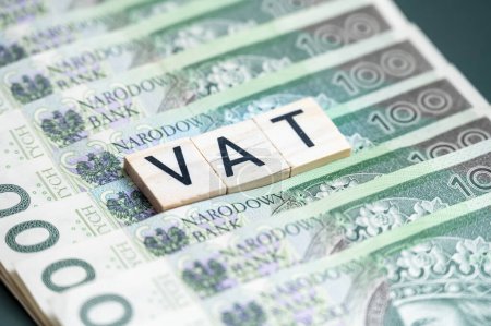 Großaufnahme von hölzernen Buchstabenblöcken mit der Schreibweise "VAT" auf einem Stapel polnischer Zloty-Banknoten, die Finanz- und Steuerkonzepte vermitteln.