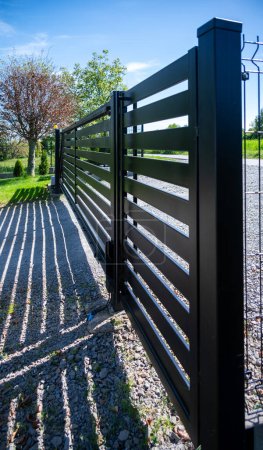 A sleek, contemporary black metal gate stands along a country road, its horizontal slats casting elongated shadows on the gravel beneath. Sunlit, vibrant greenery frames the scene.