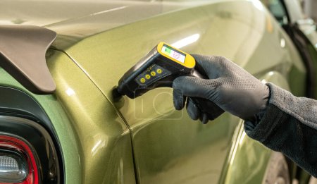 Un technicien automobile professionnel utilise une jauge d'épaisseur numérique pour mesurer l'épaisseur de la peinture sur une voiture verte moderne, assurant qualité et uniformité.