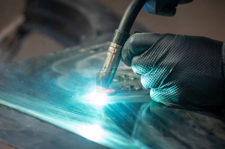 A detailed image capturing a professional welder using TIG welding technique on a metal surface. Gloved hand and blue sparks are visible.