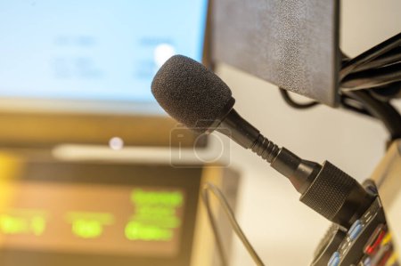 A close-up of a microphone in a broadcast studio, featuring blurred digital displays and recording equipment in the background.