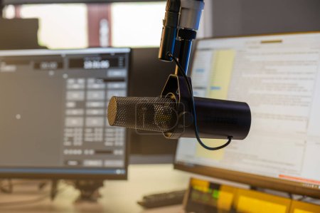 Close-up view of a high-quality microphone in a podcasting studio, with visible computer screens and documents for reference. Focus on professional audio equipment for broadcasting.
