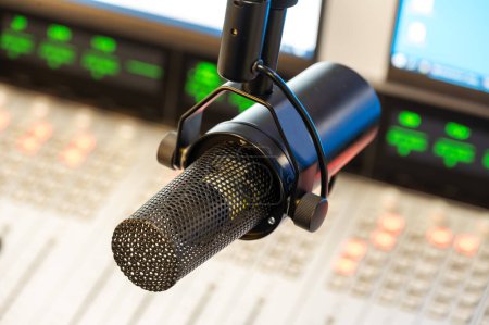 Close-up image of a professional microphone with attached headset in a broadcasting studio. The equipment is set against a background with a sound mixer and illuminated displays.