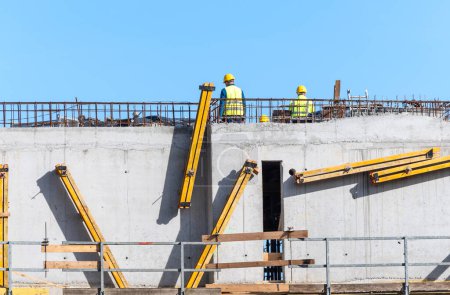 Two construction workers wearing safety gear inspecting the progress on a high concrete structure at a construction site.