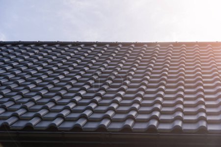 Close-up angle of a contemporary black tiled roof capturing the detailed texture and curves under a bright sun flare, emphasizing modern architectural design in residential housing.