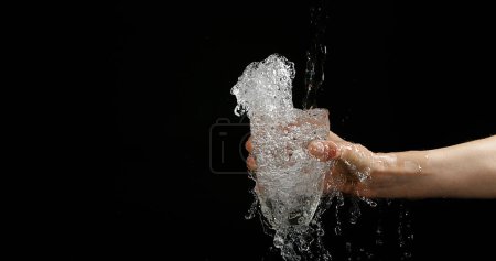 Téléchargez les photos : Les mains de la femme et l'eau coulant dans le verre sur fond noir - en image libre de droit