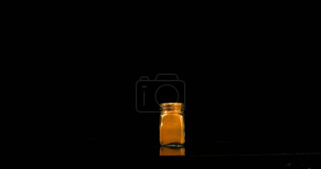 Foto de Cúrcuma, curcuma longa, Polvo en un frasco pequeño que cae contra el fondo negro, Especia india - Imagen libre de derechos