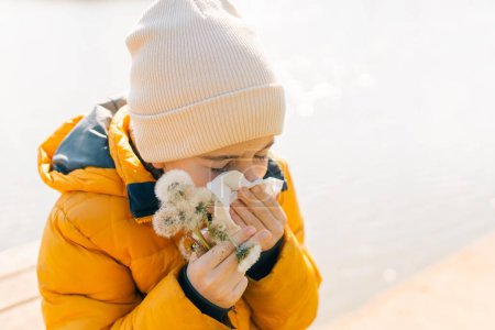  Ein kleiner Junge, 10 Jahre alt, leidet an saisonalen Frühlingsallergien. Er wird mit einer laufenden Nase und Husten gezeigt, wahrscheinlich aufgrund von Pollen von Frühlingsblumen im Hintergrund. Hochwertiges Foto