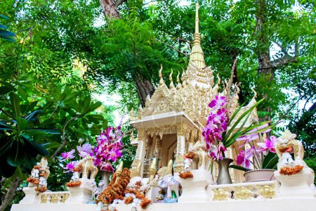 A beautiful Thai spirit house adorned with flowers and offerings, surrounded by lush greenery in a tranquil garden