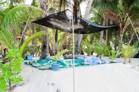 Tropical beach setup with shaded area, blankets, and cushions under palm trees in a serene environment