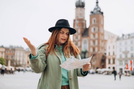 Téléchargez les photos : Triste jeune touriste féminine est perdu dans la nouvelle ville. Fille rousse marchant dans le parc et tenant une carte en papier par une journée ensoleillée à Cracovie. - en image libre de droit