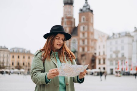 Téléchargez les photos : Attrayant jeune touriste féminine explore la nouvelle ville. Fille rousse marchant et tenant une carte papier à Cracovie. Voyager en Europe au printemps. Concept de mode de vie urbain - en image libre de droit