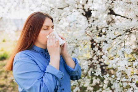 Photo for Sneezing young redhead woman with nose wiper among blooming trees in park. Portrait of sick women sneezes in white tissue, suffers from rhinitis and running nose. Symptoms of allergy. - Royalty Free Image