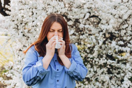 Sneezing young redhead woman with nose wiper among blooming trees in park. Portrait of sick women sneezes in white tissue, suffers from rhinitis and running nose. Symptoms of allergy.