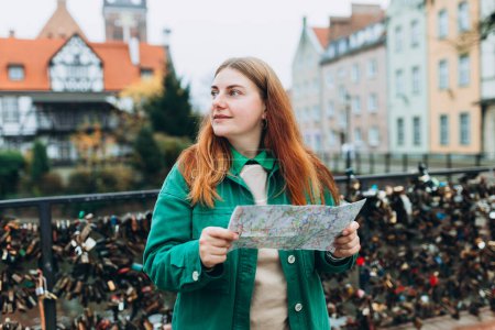 Foto de Mujer joven de 30 años con mochila sosteniendo mapa de papel contra la casa del Gremio de Molineros y el Canal Radunia en Gdansk, concepto de vacaciones. Mujer Buscando lugares en el día de otoño - Imagen libre de derechos