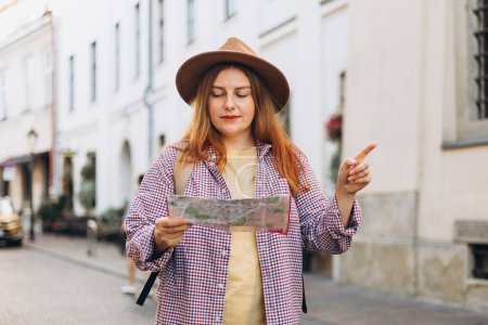 Photo for Front view of tourist examining paper map while standing on road on street near ancient building. Vacation concept. Women Searching locations and pointing finger at summer day - Royalty Free Image