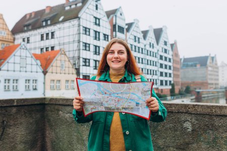 Téléchargez les photos : Attrayant jeune touriste féminine explore la nouvelle ville. Fille rousse avec sac à dos tenant une carte en papier sur la rue de la ville à Gdansk. Voyager en Europe en automne - en image libre de droit