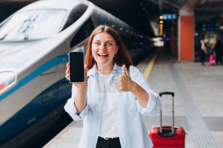 Foto de Sonriente joven mujer caucásica con maleta roja sosteniendo teléfono inteligente con pantalla en blanco en la estación de tren. maqueta en línea Wi-Fi concepto de Internet, tren de espera hipster feliz en la plataforma de la estación - Imagen libre de derechos