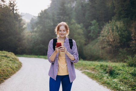 Téléchargez les photos : Fille touristique marche dans la forêt et en utilisant un smartphone. Jeune femme avec sac à dos randonnée sur la route dans la nature. Belle femme des années 30 marchant sur la nature - en image libre de droit