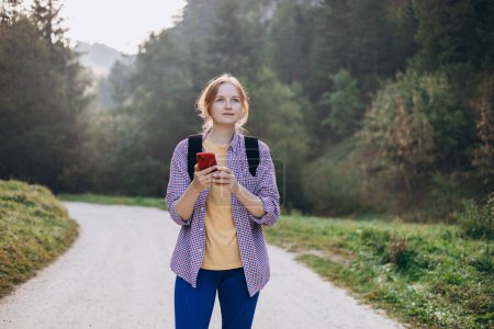 Téléchargez les photos : Fille touristique marche dans la forêt et en utilisant un smartphone. Jeune femme avec sac à dos randonnée sur la route dans la nature. Belle femme des années 30 marchant sur la nature - en image libre de droit