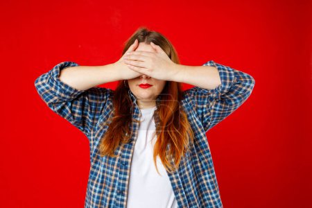 Portrait of young scared woman covering eyes with hands while standing against gray studio background. Confused girl close eyes with palms ignoring something