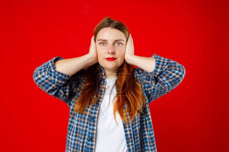 Photo for Young beautiful woman covering her ears while standing against red studio background. The concept hear no evil - Royalty Free Image