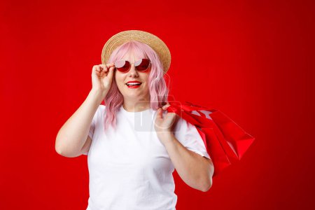 Porträt einer aufgeregten schönen Frau mit Sonnenbrille und roten Einkaufstüten vor rotem Hintergrund
