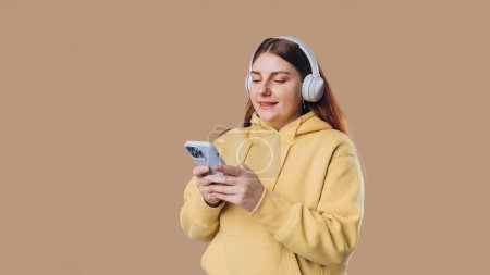Beautiful young woman wearing white wireless headphones and listening to music and using smart phone, girl isolated over colorful background wall