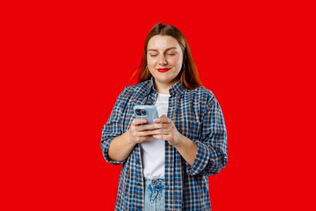 Lächelnde junge brünette Frau mit Smartphone, die isoliert auf rotem Hintergrund posiert, Studioporträt. Nette schöne junge Frau chattet per Telefon