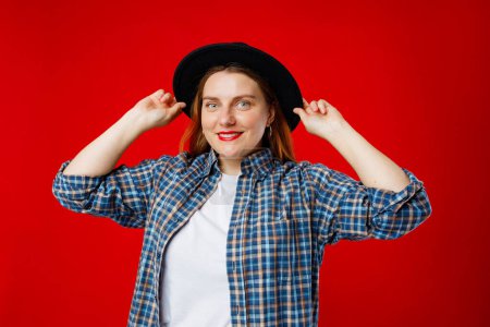 Portrait of young beautiful cute cheerful woman in hat smiling looking at camera over red background.
