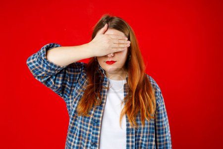 Portrait of young scared woman covering eyes with one hand while standing against red background. Confused girl close eyes with palm